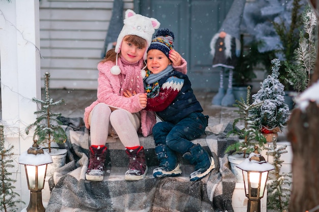 Niños felices sentados en el porche de la casa decorada de Navidad, nevando al aire libre. Feliz Año Nuevo y feliz Navidad. Invierno mágico