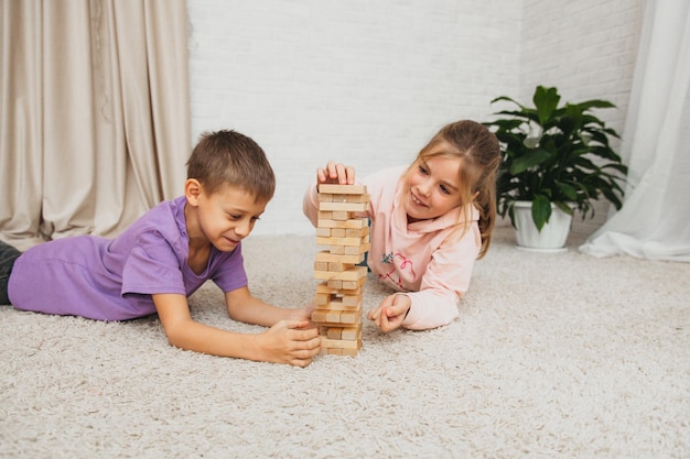 Los niños felices se sientan en el suelo y juegan un juego educativo (jenga) y se paran en una torre de bloques