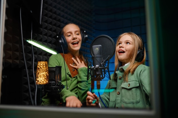 Niños felices sonrientes con auriculares cantando canciones en el estudio de grabación. Ver a través de la ventana