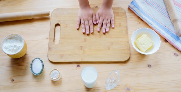 Los niños hacen masa de pastelería hecha a mano en una mesa de madera en la cocinaTabla de cortar harina rodillo toalla vaso de leche aceite de salAislamiento familiar actividad en el hogar con niñosConcepto de panadería culinaria