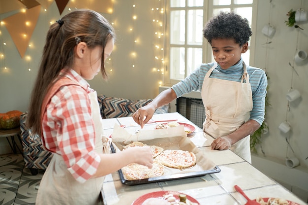 Niños haciendo pizza juntos en la mesa, agregando ingredientes a la masa.