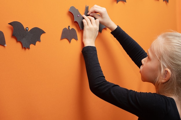 Niños de Halloween. Chica colgando murciélago cortado en papel negro hecho en casa sobre fondo naranja, espacio de copia, primer plano. Estado de ánimo de la temporada de Halloween. Concepto de decoración del hogar de Halloween