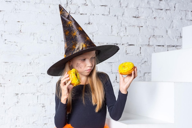 Niños de Halloween Una niña rubia sonriente con un disfraz de bruja sosteniendo donuts amarillos en sus manos sentada en los escalones