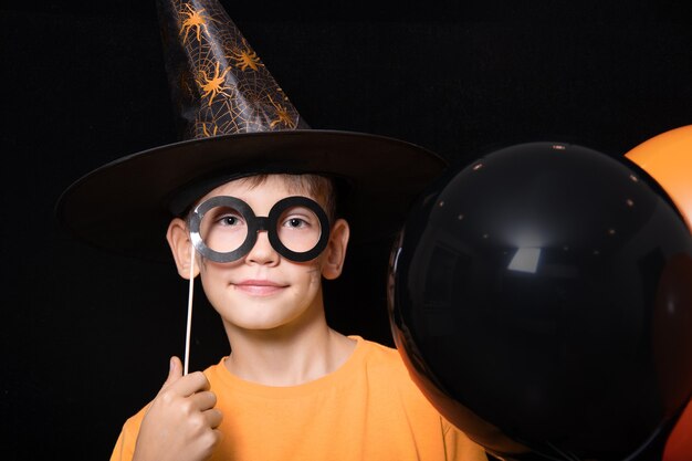 Niños de Halloween. Un niño con sombrero de mago y con una máscara de gafas negras sosteniendo globos naranjas y negros sobre un fondo negro. Listo para el truco o trato navideño.
