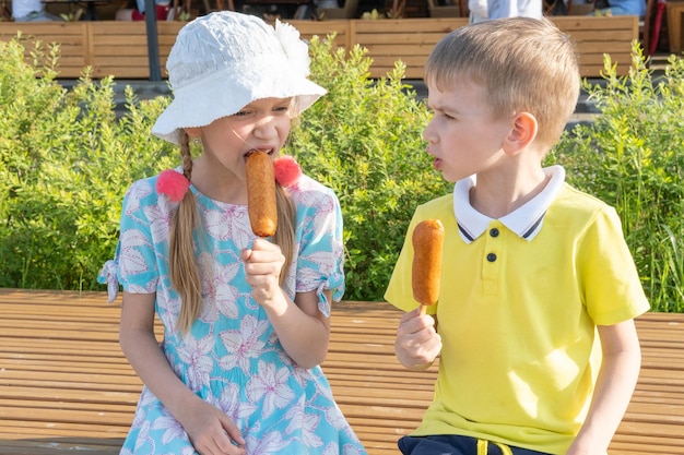 Niños hambrientos comiendo comida callejera sentados en un banco del parque Un niño y una niña comiendo salchichas fritas en un palo