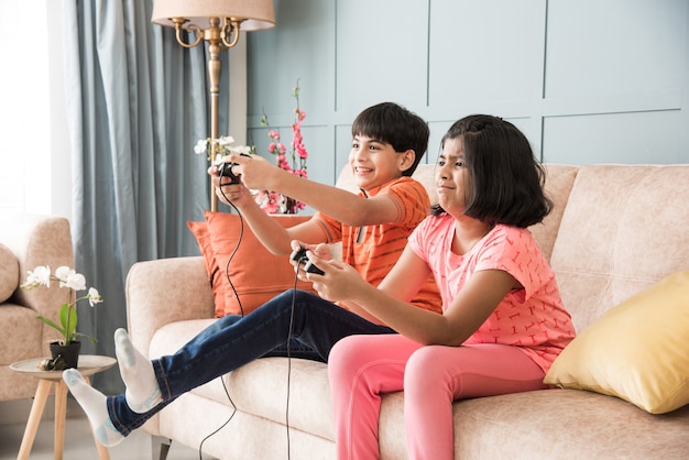 Niños indios asiáticos alegres jugando videojuegos con controlador o joystick, sentados en un sofá, sofá