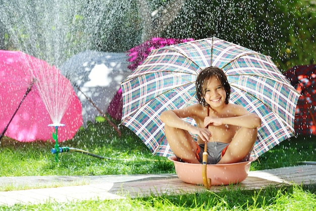 Niños jugando con agua de riego sosteniendo paraguas en patio trasero de verano