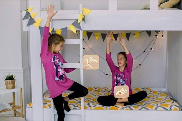 Niños jugando en el dormitorio. Dos divertidas niñas felices de 10 años, hermanas en pijamas coloridos, divirtiéndose en una litera, sosteniendo lámparas de noche de madera.
