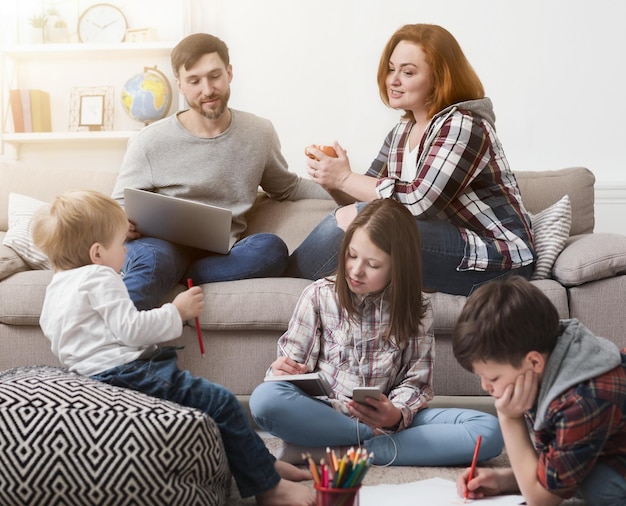 Foto niños jugando y usando dispositivos en el piso mientras los padres se relajan con la computadora portátil en el sofá