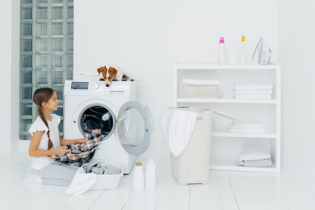 Niños de limpieza y concepto de tareas domésticas Niño feliz descarga lavadora pone ropa limpia lavada en el lavabo perro curioso mira desde arriba yace en la lavadora en el cuarto de lavado en casa