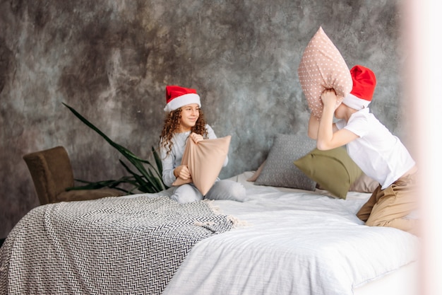 Niños lindos entre adolescentes con sombreros de Santa y pijama jugando en la cama con la almohada, la mañana de Navidad, fiesta infantil