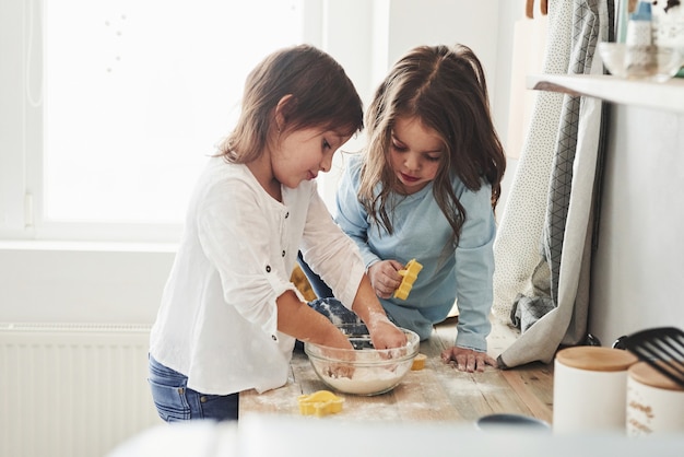 Niños lindos. Amigos de preescolar que aprenden a cocinar con harina en la cocina blanca.