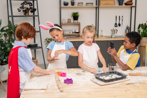 Niños lindos felices mirando galletas crudas hechas a sí mismas mientras están de pie junto a la mesa