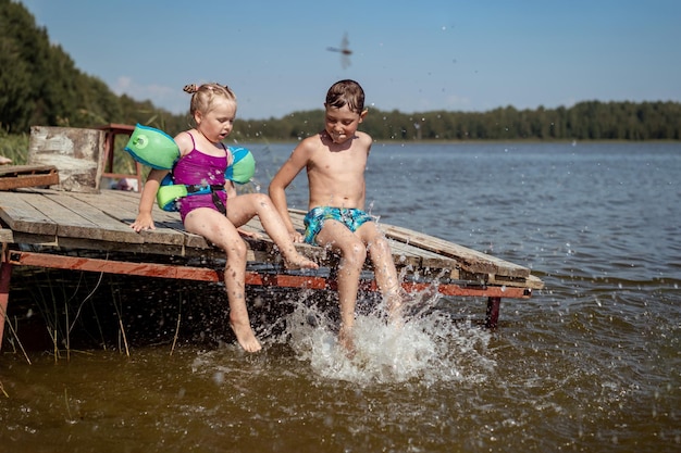 Niños lindos salpicando agua sentados en un muelle de madera junto al lago en el campo