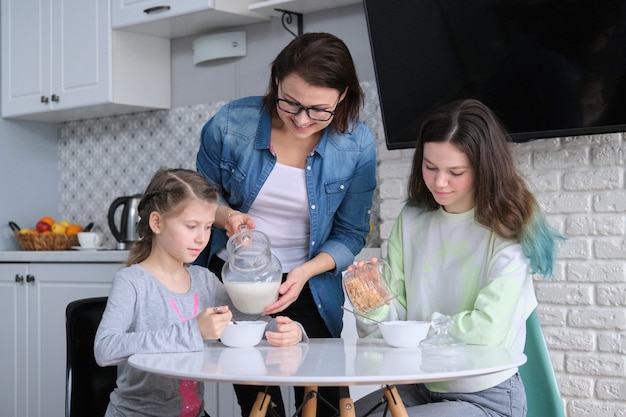 Niños con madre comiendo en casa en la cocina, dos niñas sentadas a la mesa con platos de copos de maíz y leche. Familia, alimentación, comunicación, concepto de salud.