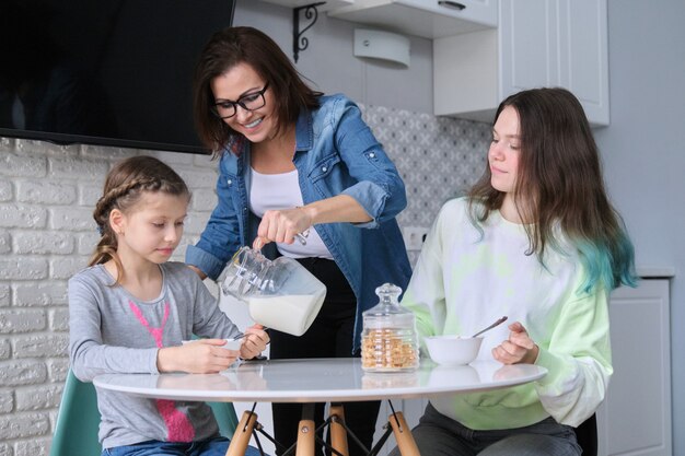Los niños con la madre comiendo en casa en la cocina, dos niñas sentadas a la mesa con platos de copos de maíz y leche