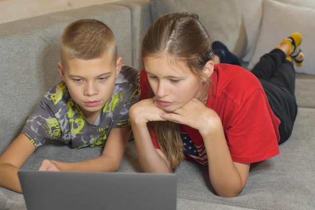 Niños mirando una computadora portátil mientras están acostados en el sofá en casa. hermano y hermana en entrenamiento remoto en línea. los niños chatean en línea con amigos