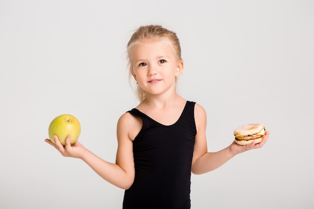 Los niños niña sonriente sostiene una manzana y una hamburguesa. Elegir comida saludable, no comida rápida, espacio para texto