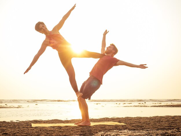 Los niños y niñas de la joven pareja deportiva están practicando ejercicios de acroyoga en la puesta de sol en la playa.