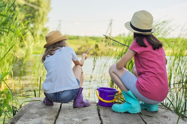 Niños niñas jugando juntos en el lago, sentados en el muelle de madera, atrapando caracoles de agua en un balde. Infancia, verano, naturaleza, concepto de niños.