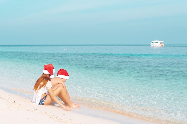 Niños, un niño y una niña con trajes de baño y sombreros de Santa, se paran en la orilla del océano en las Maldivas, el concepto de vacaciones y viajes en invierno y encuentros navideños en los trópicos