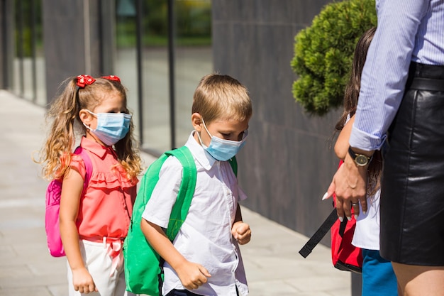 Los niños se paran con máscaras protectoras y mochilas frente a la entrada de la escuela El maestro verifica que los estudiantes cumplan con las reglas de seguridad