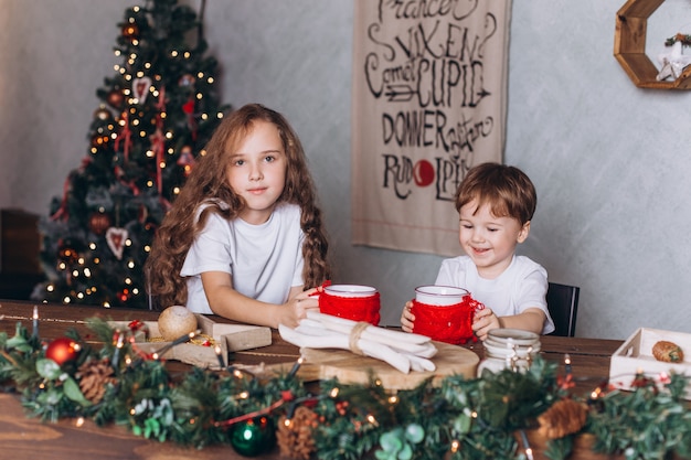 Niños pequeños en decoración navideña con té en un hogar acogedor con coloridas luces de año nuevo