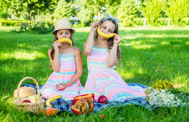 Los niños pequeños divertidos hacen emoticonos sonrientes tristes y felices con fruta de plátano en un picnic en la hierba verde en el paisaje soleado de verano, divertido.