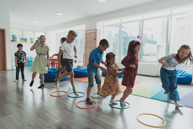 Niños pequeños de la guardería con una maestra en el suelo en el interior del aula haciendo ejercicio Saltando sobre la pista de círculos de hula hoop en el suelo