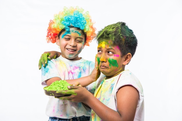 Niños pequeños indios o amigos o hermanos celebrando el festival de Holi con gulal o color en polvo, dulces, pichkari o spray, aislado sobre fondo blanco.