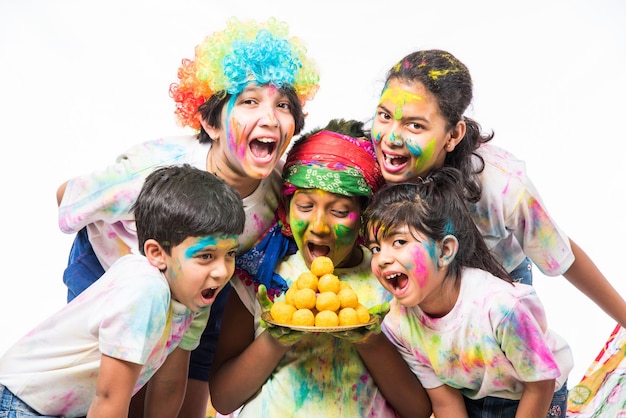 Niños pequeños indios o amigos o hermanos celebrando el festival de Holi con gulal o color en polvo, dulces, pichkari o spray, aislado sobre fondo blanco.