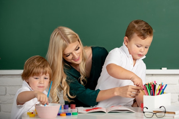 Niños pequeños con maestra de jardín de infantes dibujando en la mesa adentro aprendiendo y jugando al maestro tuto