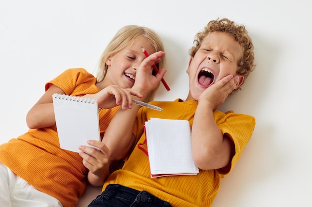 Los niños pequeños yacen en el suelo con blocs de notas y lápices aislados de fondo sin alterar