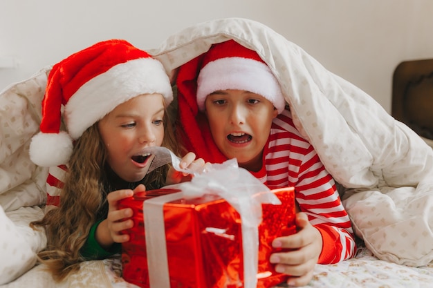 Niños en pijama y gorros navideños en la cama con regalos. concepto de navidad y año nuevo