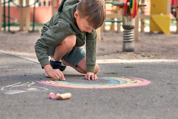 Los niños pintan al aire libre. Retrato de un niño niño dibujando una tiza de colores del arco iris sobre el asfalto en un día soleado de verano. Los niños juegan en el patio de recreo. Actividad al aire libre