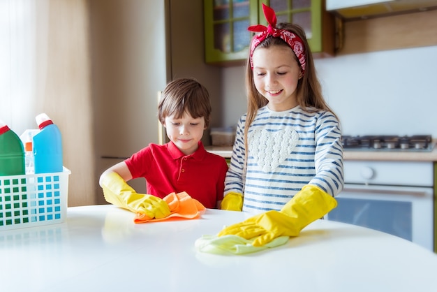 Foto los niños positivos hacen que la limpieza de la mesa de la cocina. niño y niña sonrientes frotan el polvo en la casa. concepto de limpieza