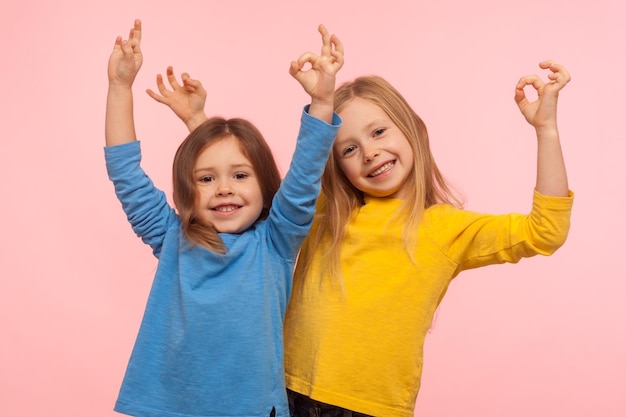 Niños que muestran que todo está bien Dos encantadoras niñas felices gesticulando bien con los dedos y sonriendo juntas, lo que significa que está bien, estamos de acuerdo en una foto de estudio interior aislada en un fondo rosa