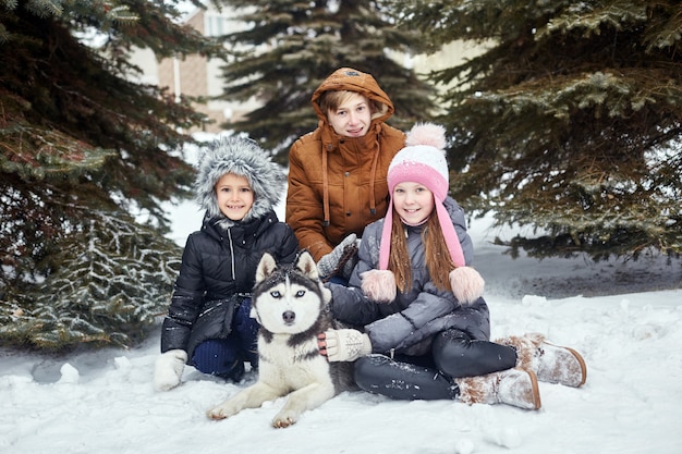 Los niños se sientan en la nieve y acariciaron al perro husky. Los niños salen a jugar con el perro husky en invierno. Caminar en el parque