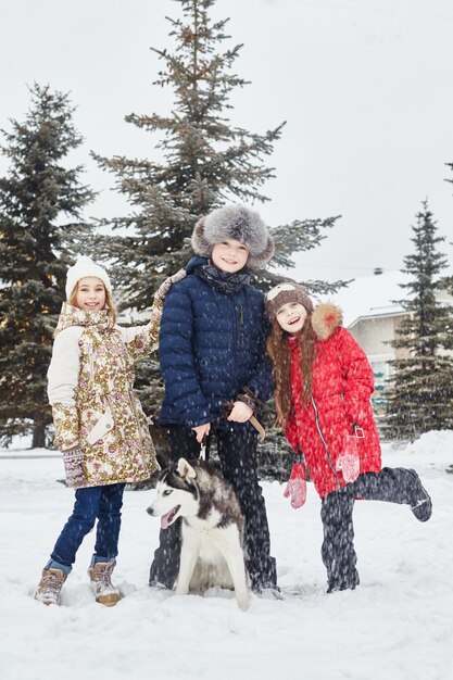 Los niños se sientan en la nieve y acariciaron al perro husky. Los niños salen a jugar con el perro husky en invierno. Pasee por el parque en invierno, alegría y diversión, perro husky con ojos azules.