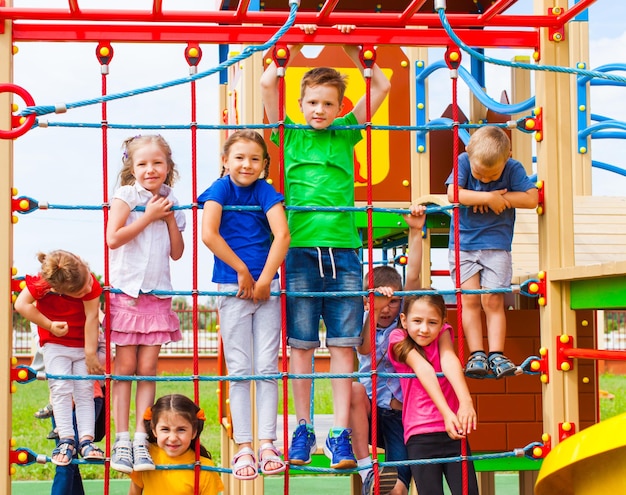 Niños sonrientes colgando de una cuerda de red colorida usando sus manos. Los niños pasan el tiempo libre haciendo ejercicio, escalando en el patio de recreo al aire libre en verano