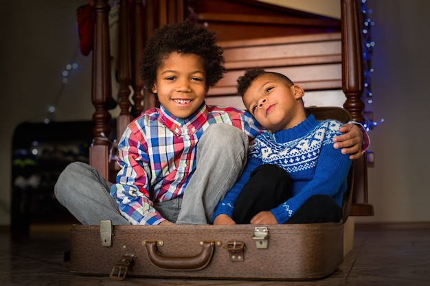 Los niños sonrientes se sientan dentro de la maleta. Chicos afro alegres junto a la escalera. Feliz de tener un hermano. Cuida a tus seres queridos.