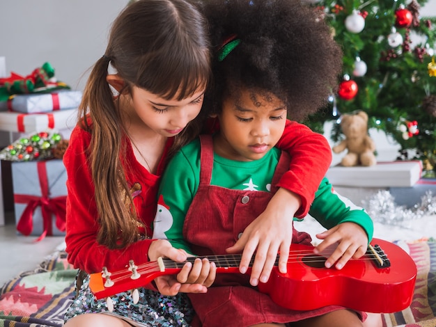 Los niños tocan la guitarra alegremente el día de Navidad con el árbol de navidad