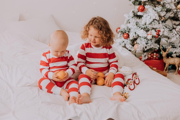 Los niños con trajes rojos y blancos comen mandarinas en la cama. hermano y hermana celebrando la Navidad. niño y una niña están jugando en casa. estilo de vida. espacio para texto. foto de alta calidad