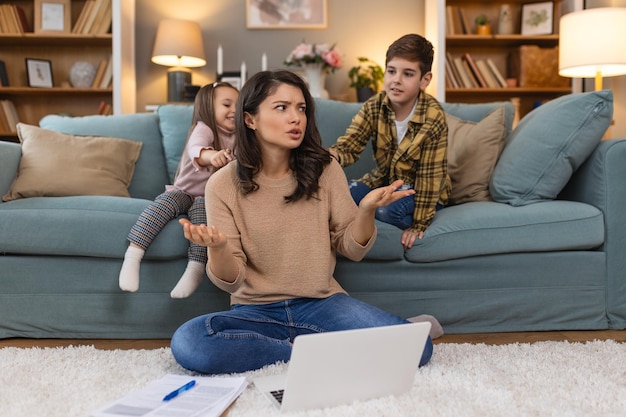 Niños traviesos pequeños haciendo ruido y distraendo a la madre independiente tratando de concentrarse en la computadora portátil y apretando la cabeza trabajando en la mesa en la sala de estar ligera