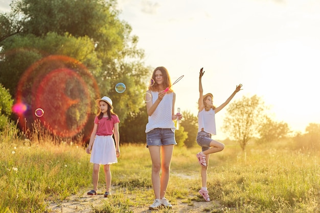 Los niños tres niñas jugando con pompas de jabón, el adolescente sopla a los más jóvenes atrapan burbujas. Hermoso paisaje natural, fondo de pradera de verano, infancia feliz