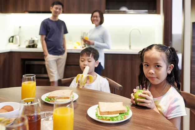 Los niños vietnamitas preadolescentes sentados en la mesa de la cocina y comiendo sándwiches y frutas para el desayuno