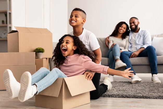 No apartamento novo. feliz família afro-americana se divertindo e comemorando o dia da mudança, irmão animado montando sua irmã em caixa de papelão, pais abraçando enquanto estão sentados no sofá ao fundo