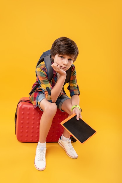 No azul. Menino adorável entediado segurando seu tablet enquanto está sentado em sua bagagem