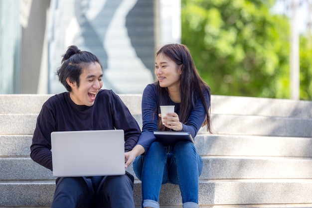 No campus, um lindo casal em trajes casuais usa um laptop enquanto está sentado na escada externa.