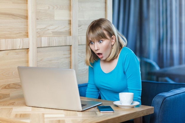 ¡No creas! El retrato de vista lateral de una joven empresaria conmocionada emocionalmente con una camiseta azul está sentado en un café, leyendo noticias y trabajando de forma remota con grandes ojos sorprendidos mirando a la computadora portátil. Interior